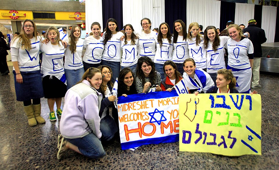 Young women in Welcome Home shirts holding Hebrew signs to greet new immigrants