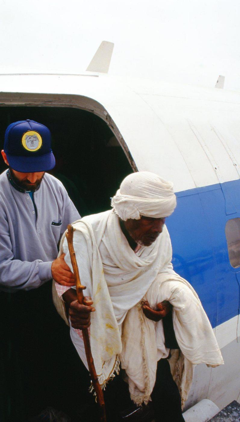 An elderly man in a white shawl, leaning on a walking stick, takes his first step down from the aircraft door, May 1991