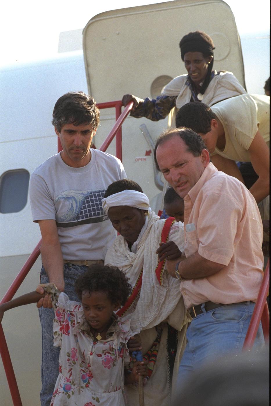 Ground crew members reach out to help a small girl down the airplane stairs, May 1991
