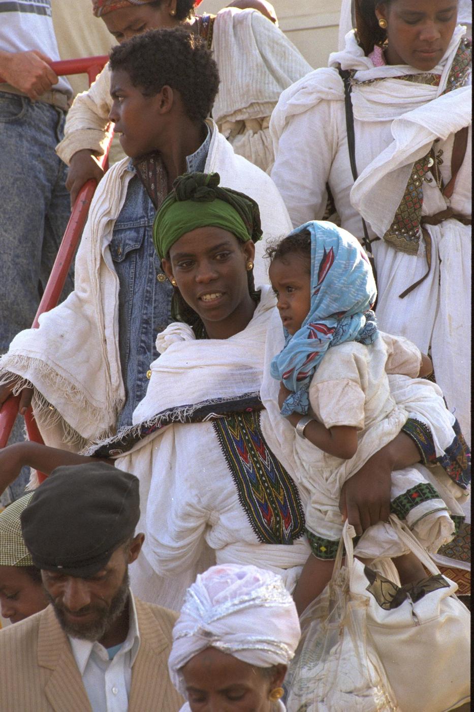 A young mother in traditional white dress, her baby wrapped against her chest, steps off a Boeing jet in Israel, May 1991